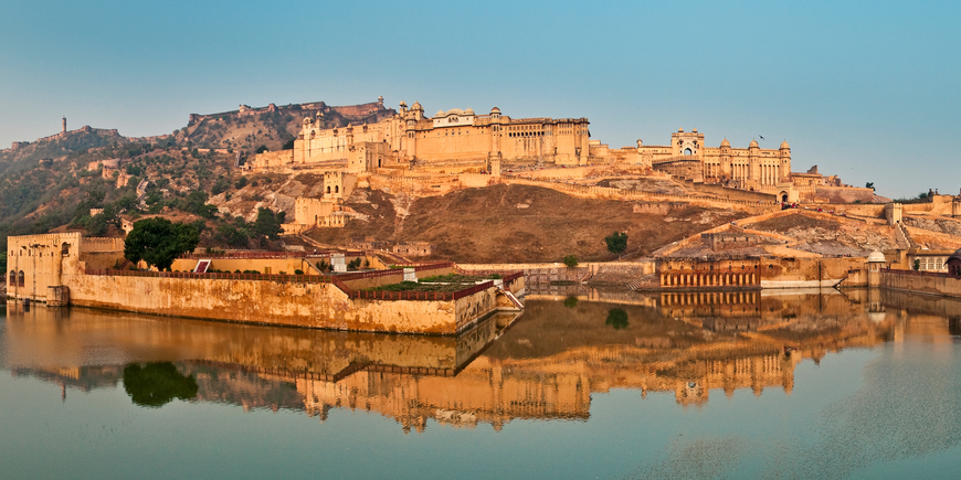 Amber Fort near Jaipur, India, rising above Maota Lake at golden hour, with its sandstone ramparts, palace terraces, and hilltop fortifications reflected in the water