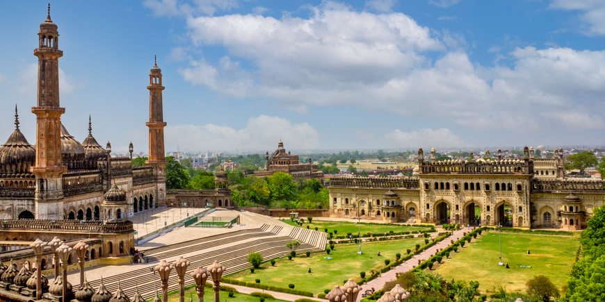 Bara Imambara complex in Lucknow, India, with its ornate gateway, towering minarets, sweeping stone staircases, and manicured Mughal-style gardens beneath a bright blue sky