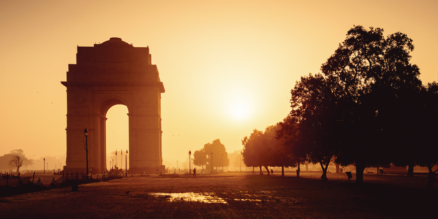 India Gate war memorial in New Delhi silhouetted against a golden sunrise, with lamp-lit lawns and mature trees in the soft morning haze