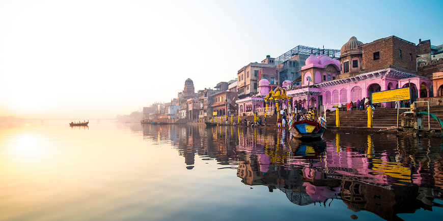 Colorful riverfront ghats of Varanasi, India, along the Ganges at sunrise, with pink-hued temples and buildings, wooden boats, and soft golden light reflected on the calm water